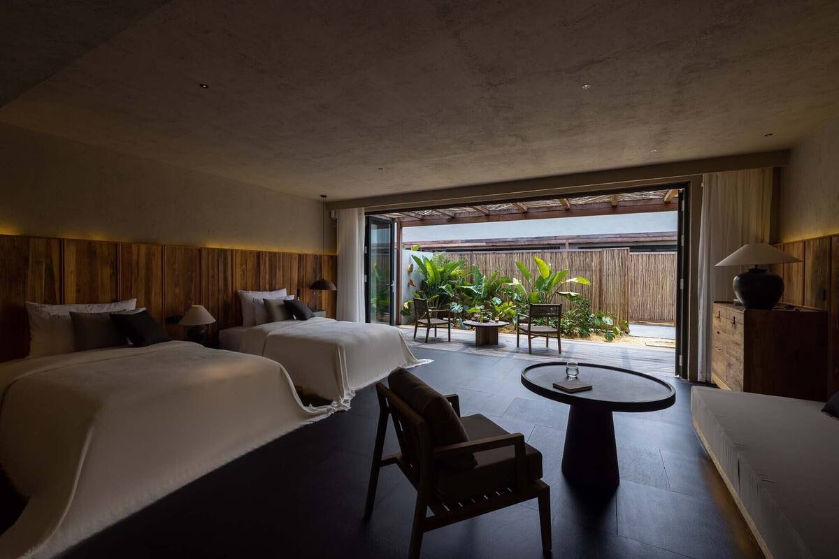 A hotel room with two beds, a small table, a chair, and a view of a garden through a glass door.