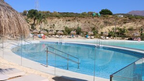 A swimming pool with sun loungers and thatched umbrellas, surrounded by a sandy beach and a hilly landscape. - Garden Apartment at Alcazaba Lagoon (Casares)