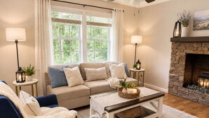 Ein Wohnzimmer mit Sofa, Sessel, Couchtisch, Kamin und zwei mit Vorhängen versehenen Fenstern.