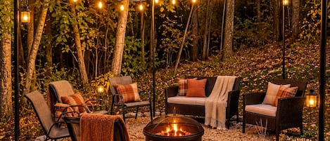 An outdoor seating area with chairs and a fire pit, surrounded by pebbles and trees, with string lights overhead.