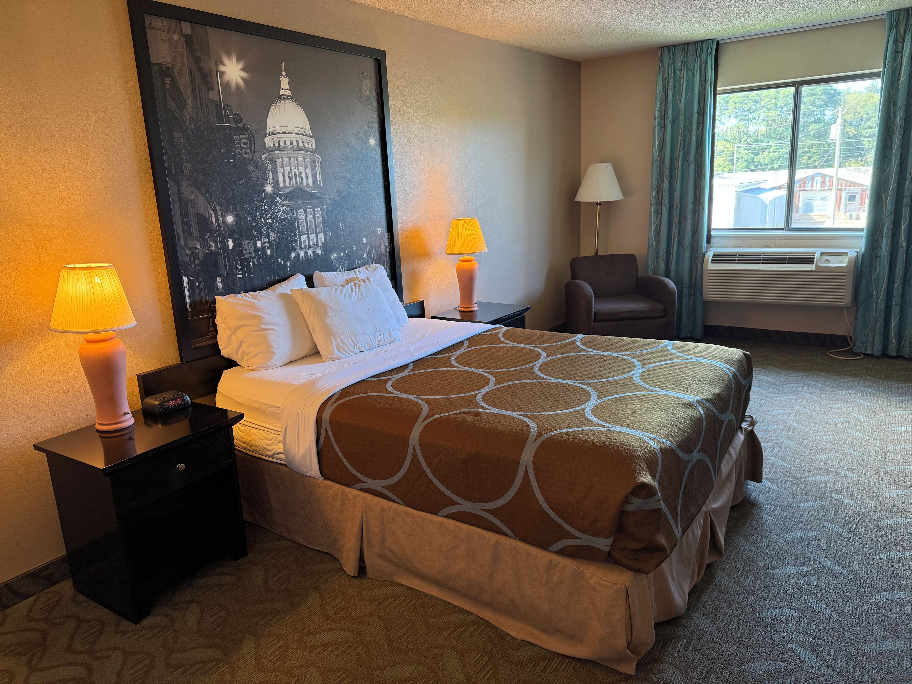 A hotel room with a bed, two bedside lamps, a chair, an air conditioning unit, and a large framed picture on the wall.
