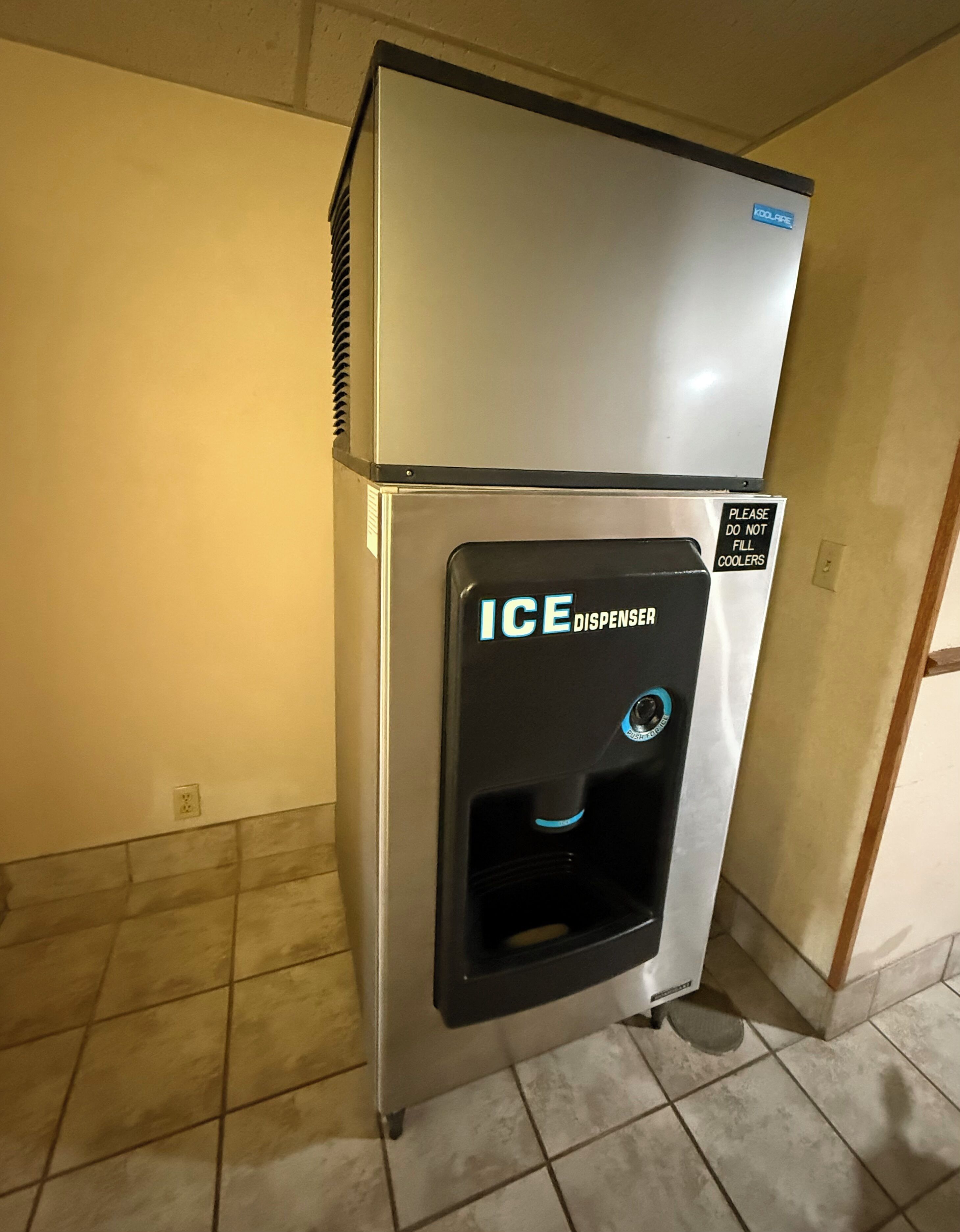 A compact ice dispenser with a black front panel and a silver top, situated in a corner with tiled flooring.