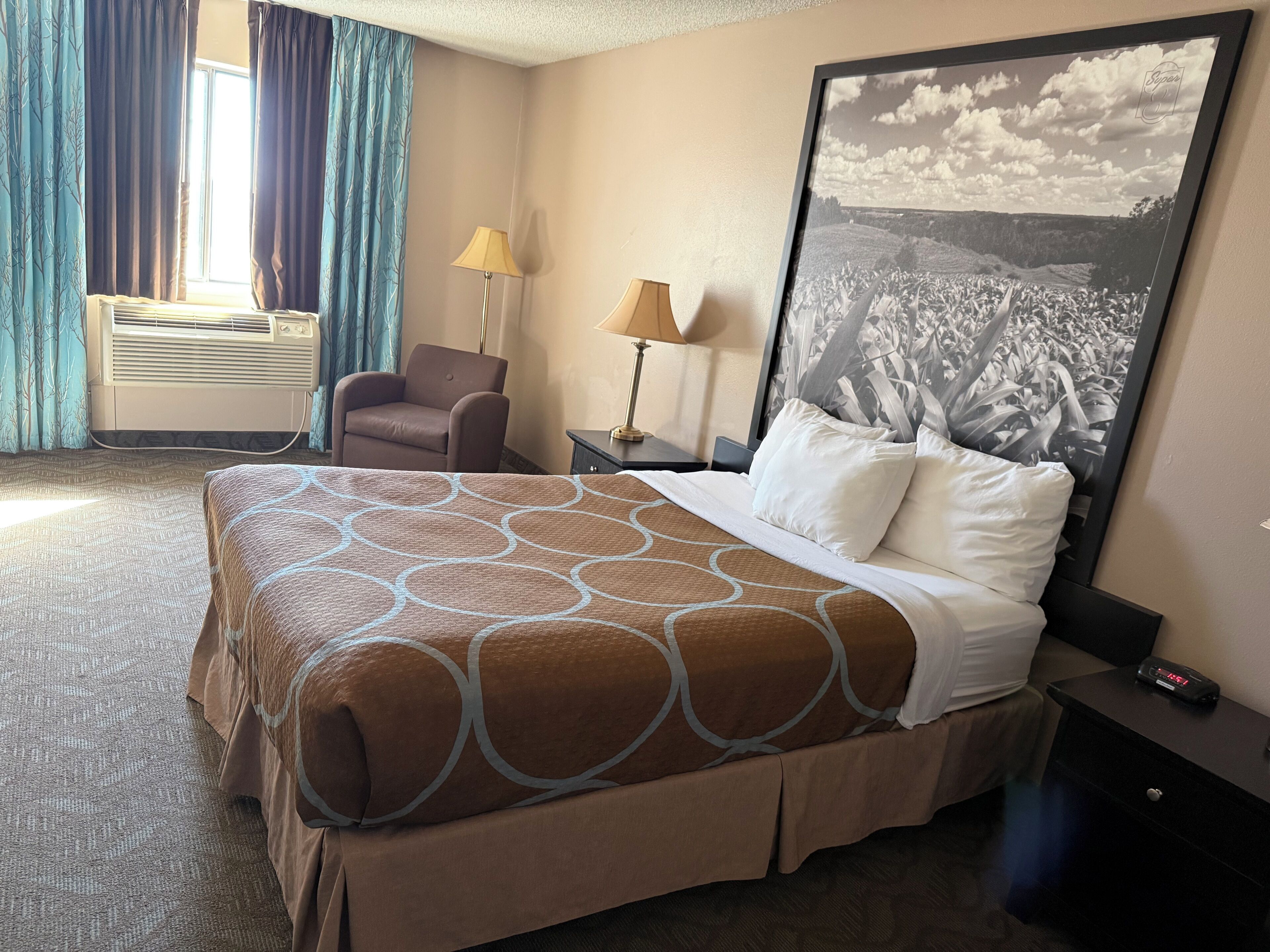 A hotel room with a large bed, a chair, a desk, and a wall-mounted air conditioner.