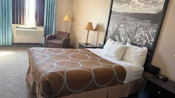 A hotel room with a large bed, a chair, a desk, and a wall-mounted air conditioner.