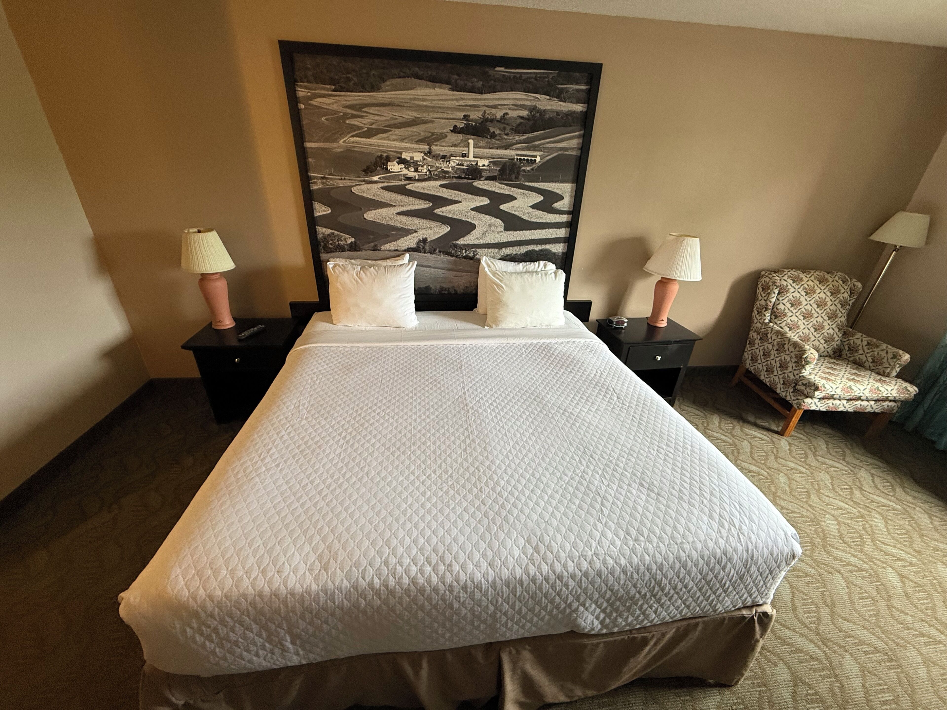 Une chambre d’hôtel avec un grand lit, deux tables de chevet avec des lampes, une chaise et un grand tableau représentant un paysage accroché au mur.