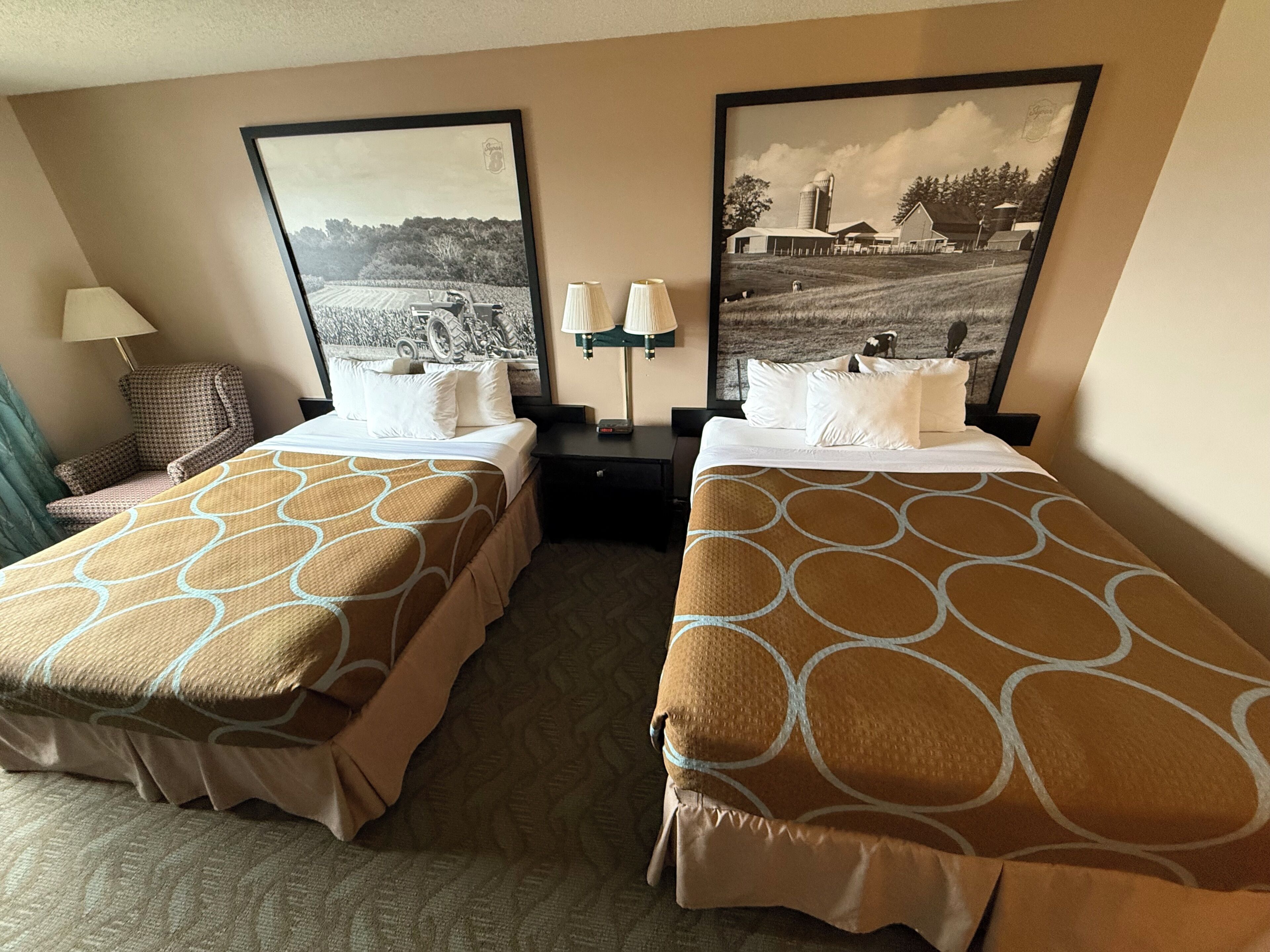 A hotel room with two beds, a chair, a lamp, and two framed pictures on the wall.