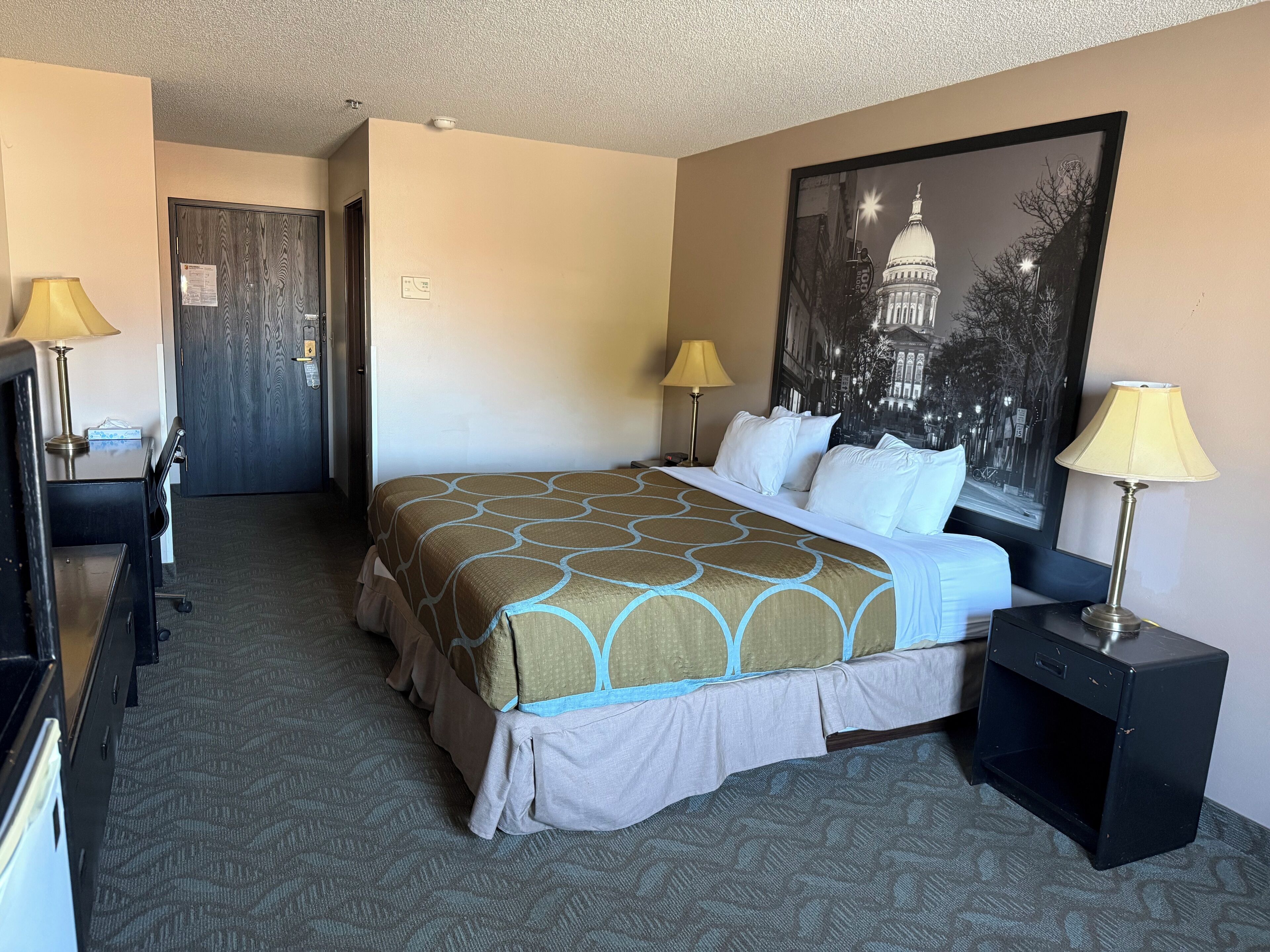 A hotel room with a bed, two bedside lamps, a desk with a lamp, a chair, a door, and a framed picture on the wall.