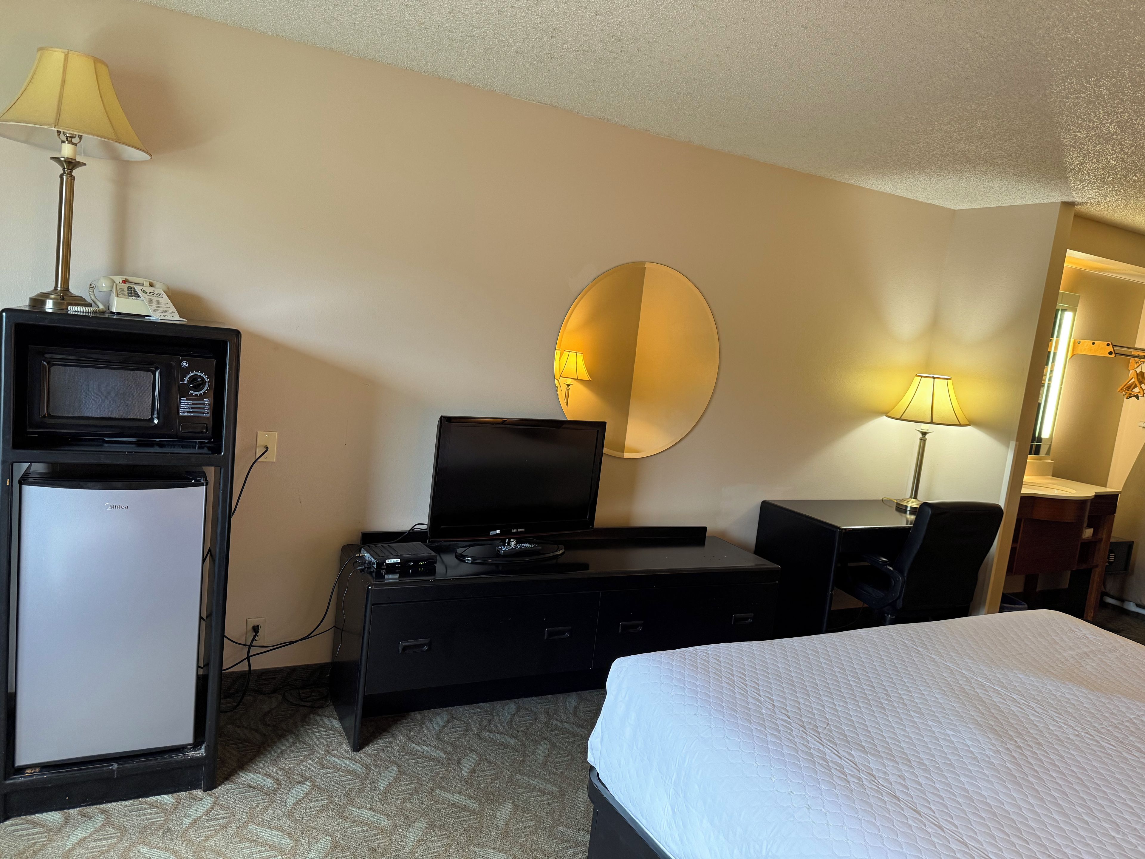 Habitación de hotel con cama, escritorio con computadora, refrigerador pequeño, microondas y espejo.