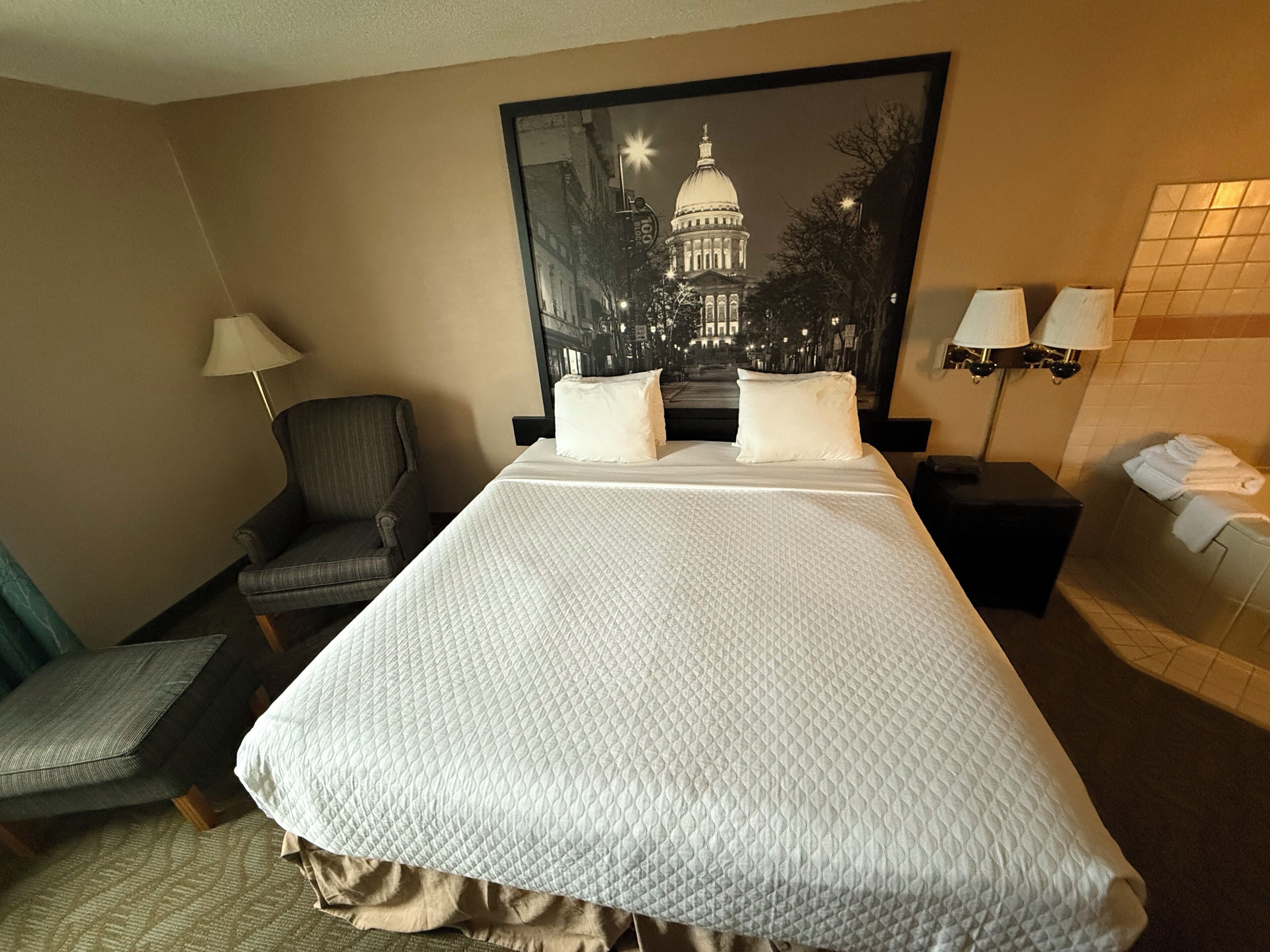 Een hotelkamer met een groot bed, een stoel, een nachtkastje, een lamp en een aan de muur bevestigd schilderij.