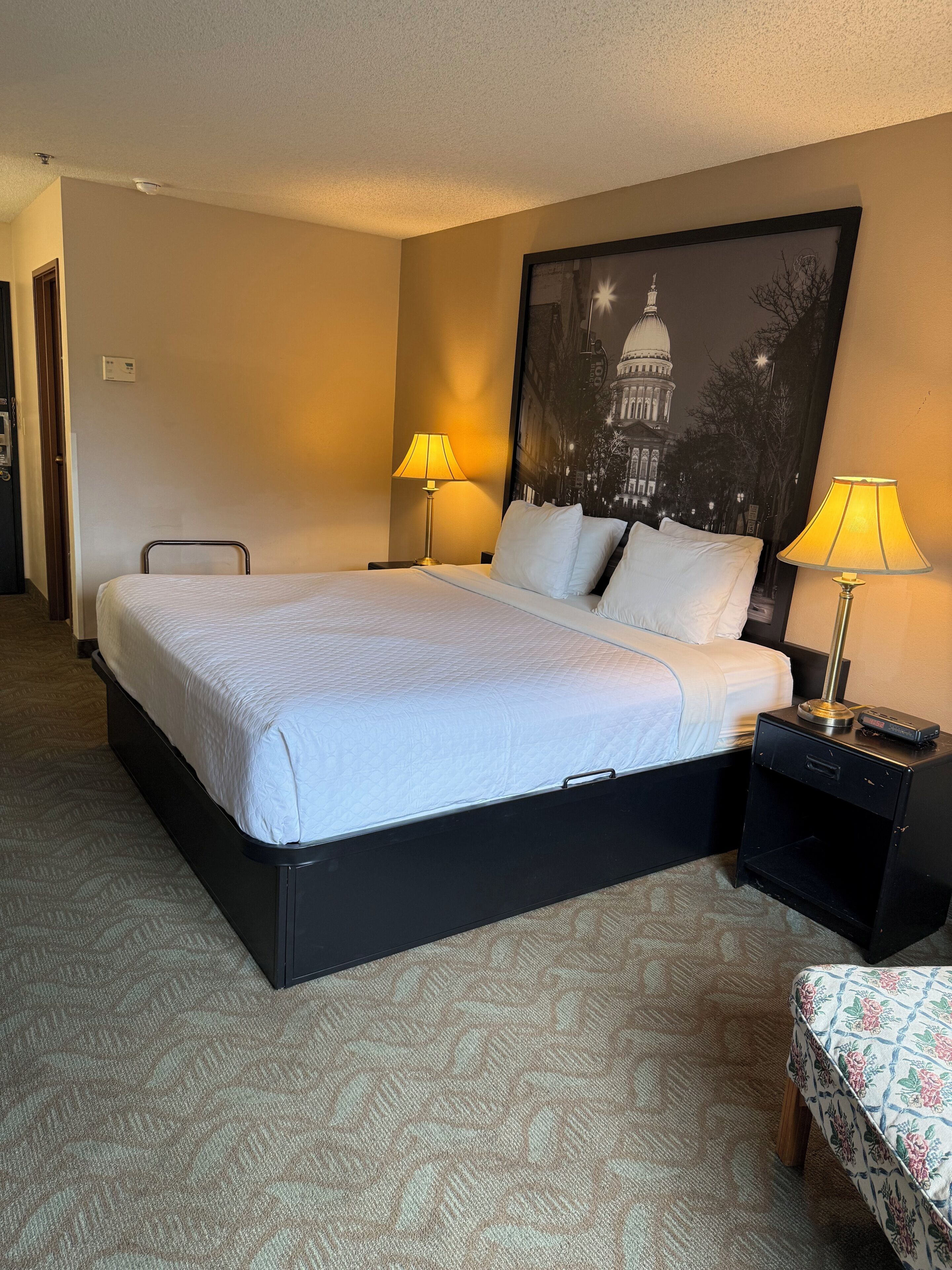 A hotel room with a bed, two bedside lamps, a nightstand, a chair, a framed picture on the wall, and a carpeted floor.