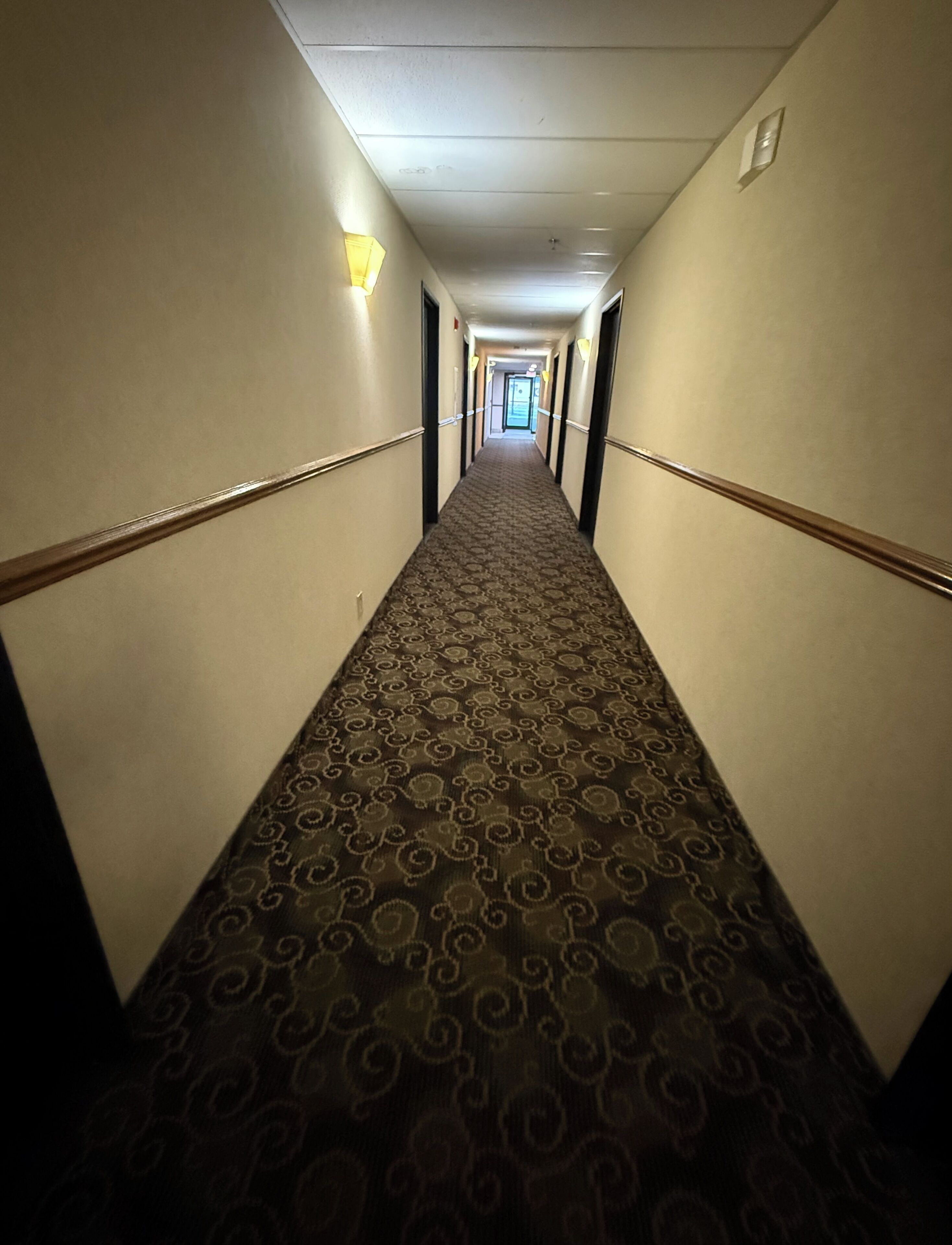 Ein Hotelgang mit Musterteppich, Wandleuchten und einer Tür am Ende.