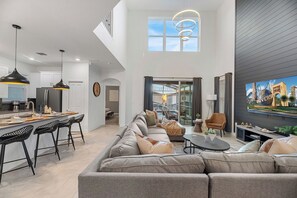 A modern living room with a large sofa, a dining area with bar stools, and a TV mounted on the wall. - FREE Pool/Hot-Tub Heat - Gated Resort Disney Home (Davenport)