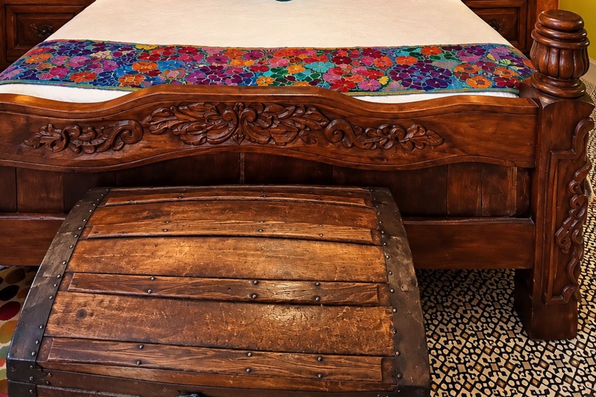 Ein hölzernes Bett mit floraler Tagesdecke und Zierkissen, neben dem Nachttische mit Lampen stehen, an einer strukturierten, orangefarbenen Wand.