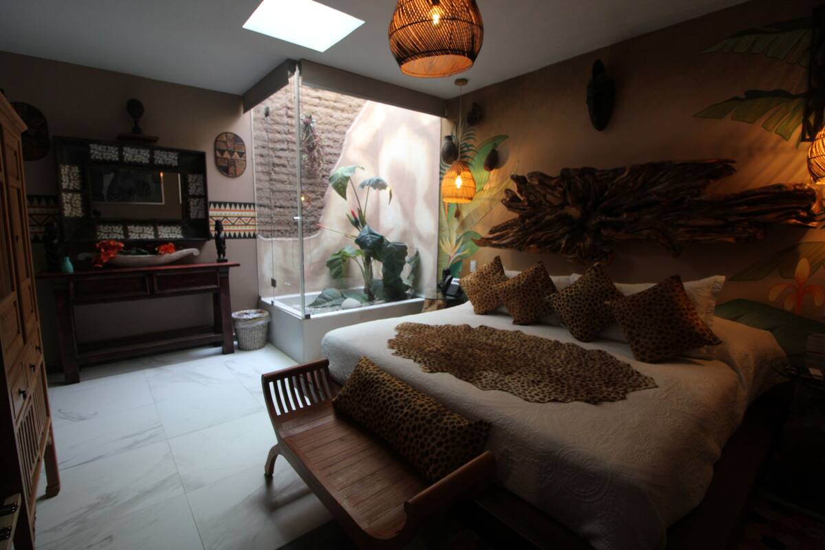 Een slaapkamer met een bed, een bankje, een bureau en een groot raam.
