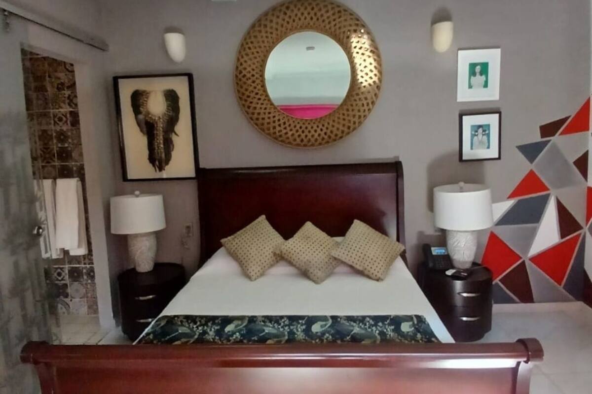 Een slaapkamer met een houten bed, een ronde spiegel, een hoofdbord met decoratieve kussens, een wandlamp en een wand met een geometrisch patroon.