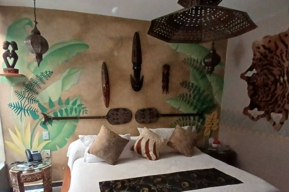 Een bed met een witte spreideken, meerdere kussens, een decoratief hoofdbord met wanddecoratie, een hanglamp en een klein nachtkastje met een telefoon.