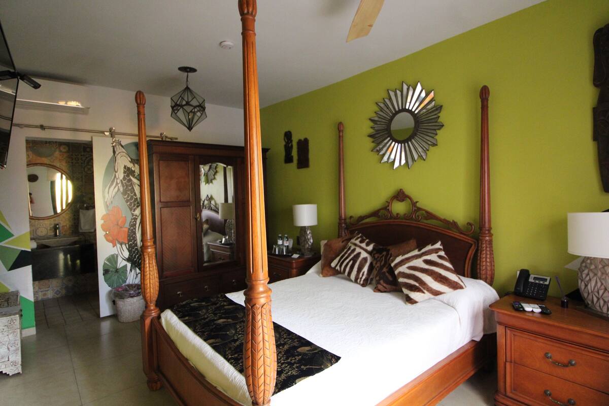 Een hemelbed met een houten hoofdbord, een spiegel aan de muur en een nachtkastje met een lamp.