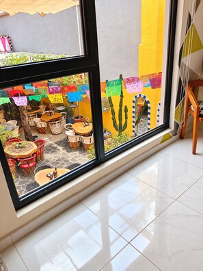 Ein Zimmer mit großem Fenster mit Blick auf eine Außenpatio-Terrasse mit Tischen und Stühlen, einem Kaktus und farbenfrohen Dekorationen.