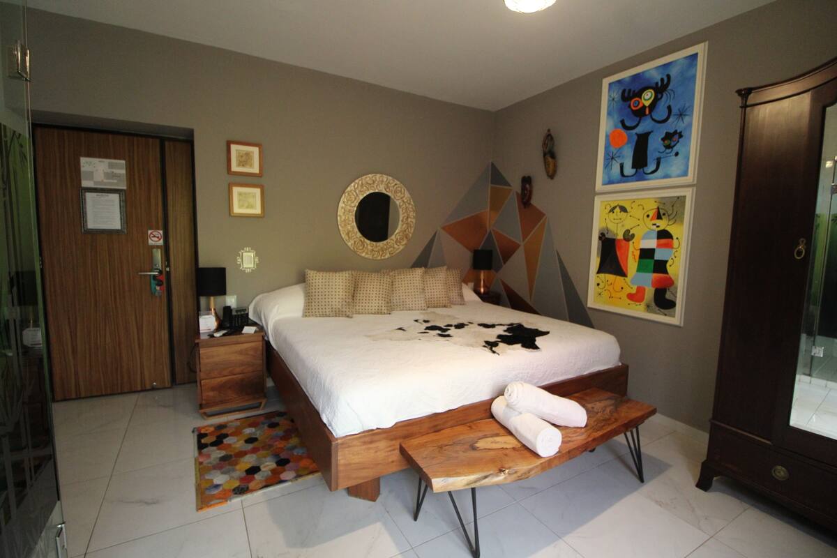 Een slaapkamer met een bed, een houten bankje, een nachtkastje, een spiegel en wanddecoratie.