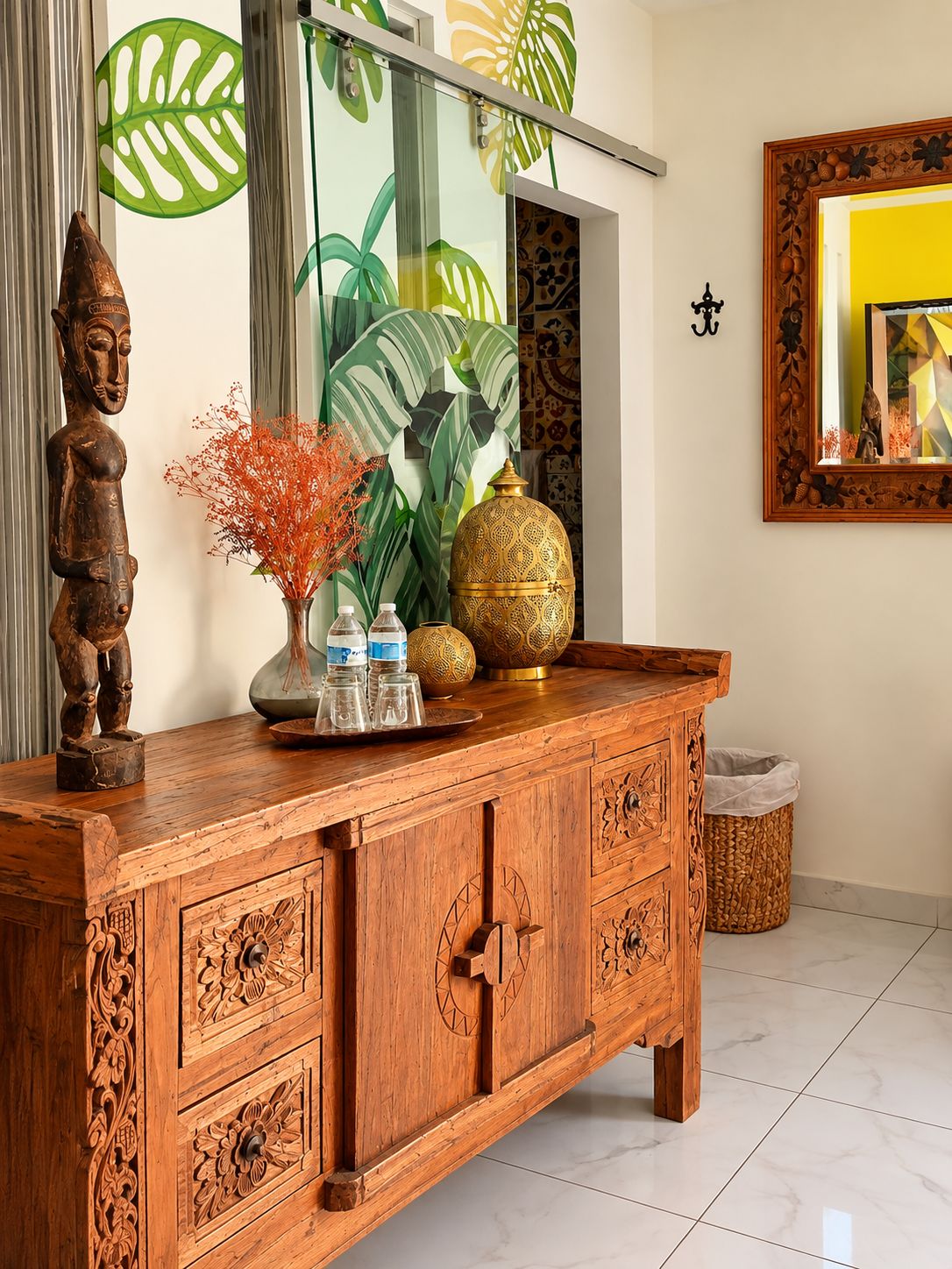 A wooden cabinet with intricate carvings, a mirror with a geometric frame, and a large plant with broad leaves.