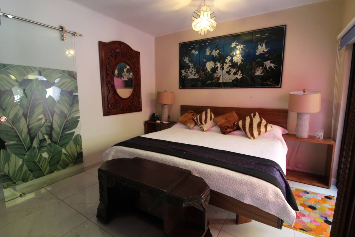 Een slaapkamer met een bed, bedlampen, een nachtkastje, een houten bankje, een spiegel, een groot bloemschilderij en een glazen deur met een plantenmuur.