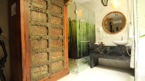 Une salle de bain dotée d’une douche à parois de verre, d’une porte en bois aux sculptures complexes, d’un miroir rond et d’un lavabo en marbre.