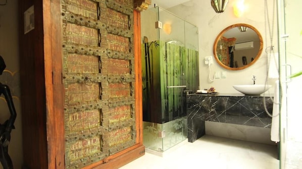 A bathroom with a glass shower enclosure, a wooden door with intricate carvings, a round mirror, and a marble sink. - Hotel Boutique Ekléctic (Guadalajara)
