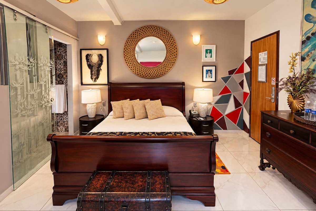 Ein Hotelzimmer mit einem großen Bett, Nachttischen, einer Holzkommode, einem Spiegel und einer geometrischen Wanddekoration.