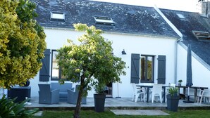 Ein weißes Gebäude mit grauen Fensterläden, ein kleiner Baum und Sitzmöglichkeiten im Freien.