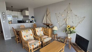 Una sala de estar con muebles de mimbre, una mesa de centro de madera, un televisor sobre un soporte de madera y una decoración de velero en la pared.