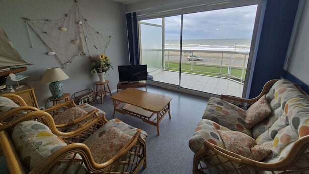 Un salón amplio con un gran ventanal con vistas a la playa, amueblado con sillas de mimbre y un sofá.