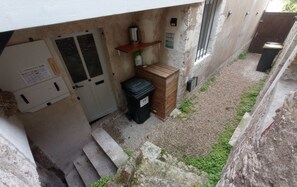 Ein schmaler Außenkorridor mit einer Tür, einem Mülleimer, einem kleinen Holfach und einem an der Wand montierten Stromverteiler.