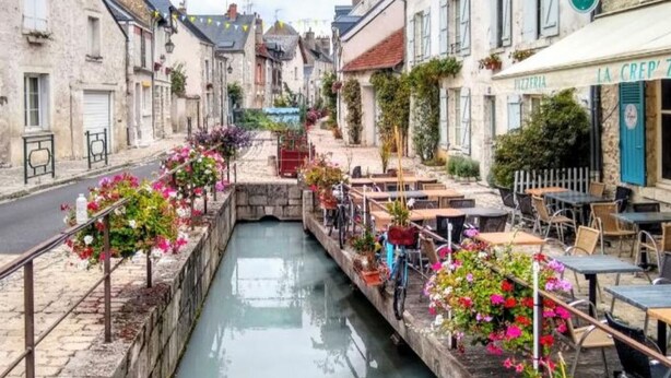 Ein schmaler Kanal mit blumenbepflanzten Ufern, Sitzgelegenheiten im Freien und Gebäuden an beiden Seiten.