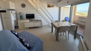Un espai vital reduït amb un sofà, una taula de menjador amb cadires, un televisor sobre un moble, una escala i una vista a la platja a través d'un gran finestral.