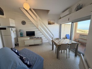 A compact living space with a sofa, a dining table with chairs, a TV on a cabinet, a staircase, and a view of the beach through a large window. - Apartment with sea view - 6 guests (Le Barcarès)
