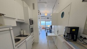 Una cuina compacta amb armaris blancs, microones, forn i cafetera. Es veu, al fons, una zona de dinar amb taula i cadira.