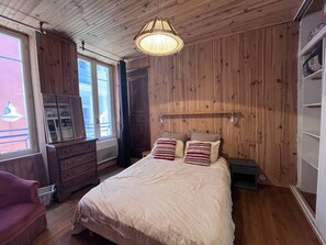 A bedroom with a wooden ceiling, a bed with striped pillows, a dresser, and a window with a view of a balcony. - Charming apartment near thermal baths - 4 guests (Cauterets)