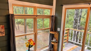 A cozy room with large windows, a wooden shelf with a cat, and a vase of flowers. - The Canopy Cabin a Nature Lovers Eclectic Treehouse (Athelstane)