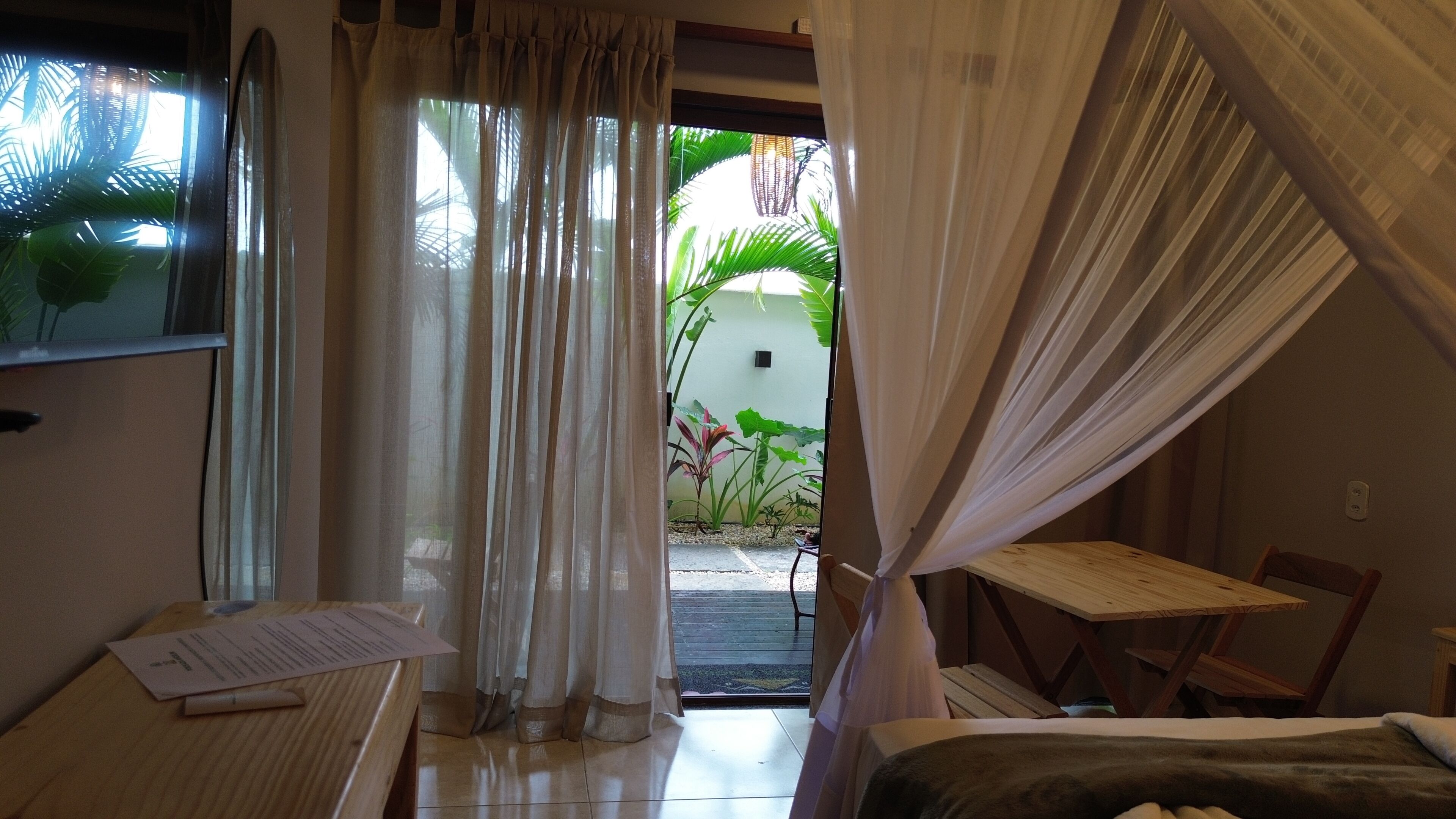 Ein Zimmer mit einem Bett, einem Schreibtisch und einem Stuhl, mit Blick auf einen Garten durch eine Tür.