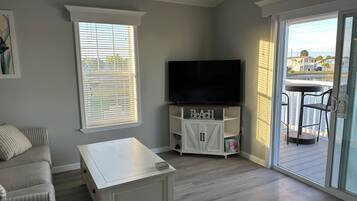 Una sala de estar con televisor, sofá, mesa y puerta corrediza de vidrio que da a un balcón con vistas al agua y casas.