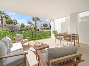 A modern outdoor living area with a sofa, chairs, and a dining table set for two, overlooking a well-maintained garden and a residential building. - Stylish 2-pax Stay in Alcazaba Lagoon (Casares)