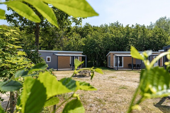 Two modern cabins with large windows, a patio table, and a grassy yard surrounded by trees. - Cosy Family Chalet With Covered Terrace (Burgh-Haamstede)