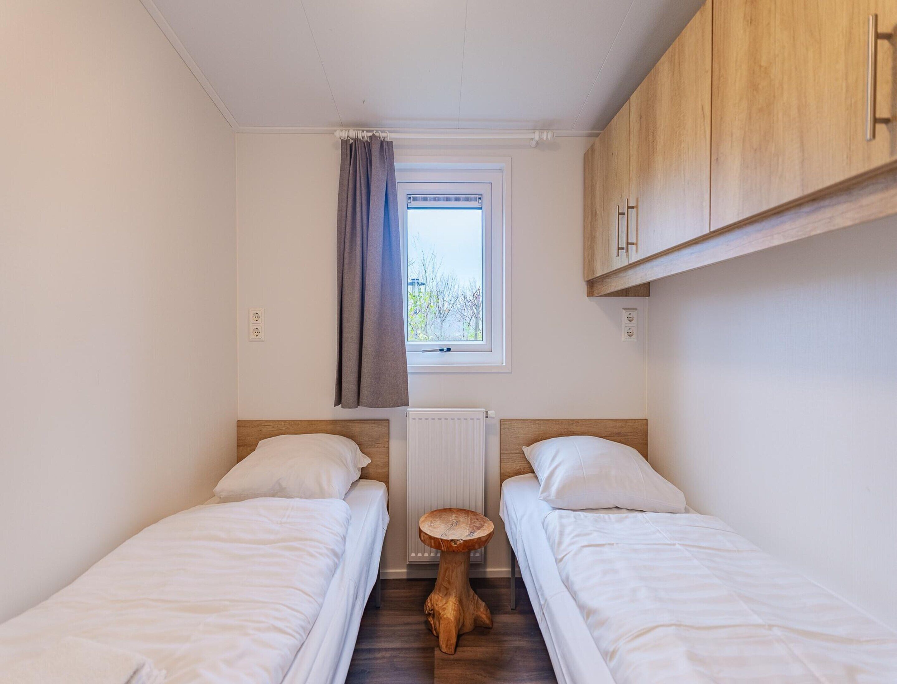 A small, well-lit room with two single beds, a wooden cabinet, a small round wooden stool, a window with a view of trees, and a white radiator.