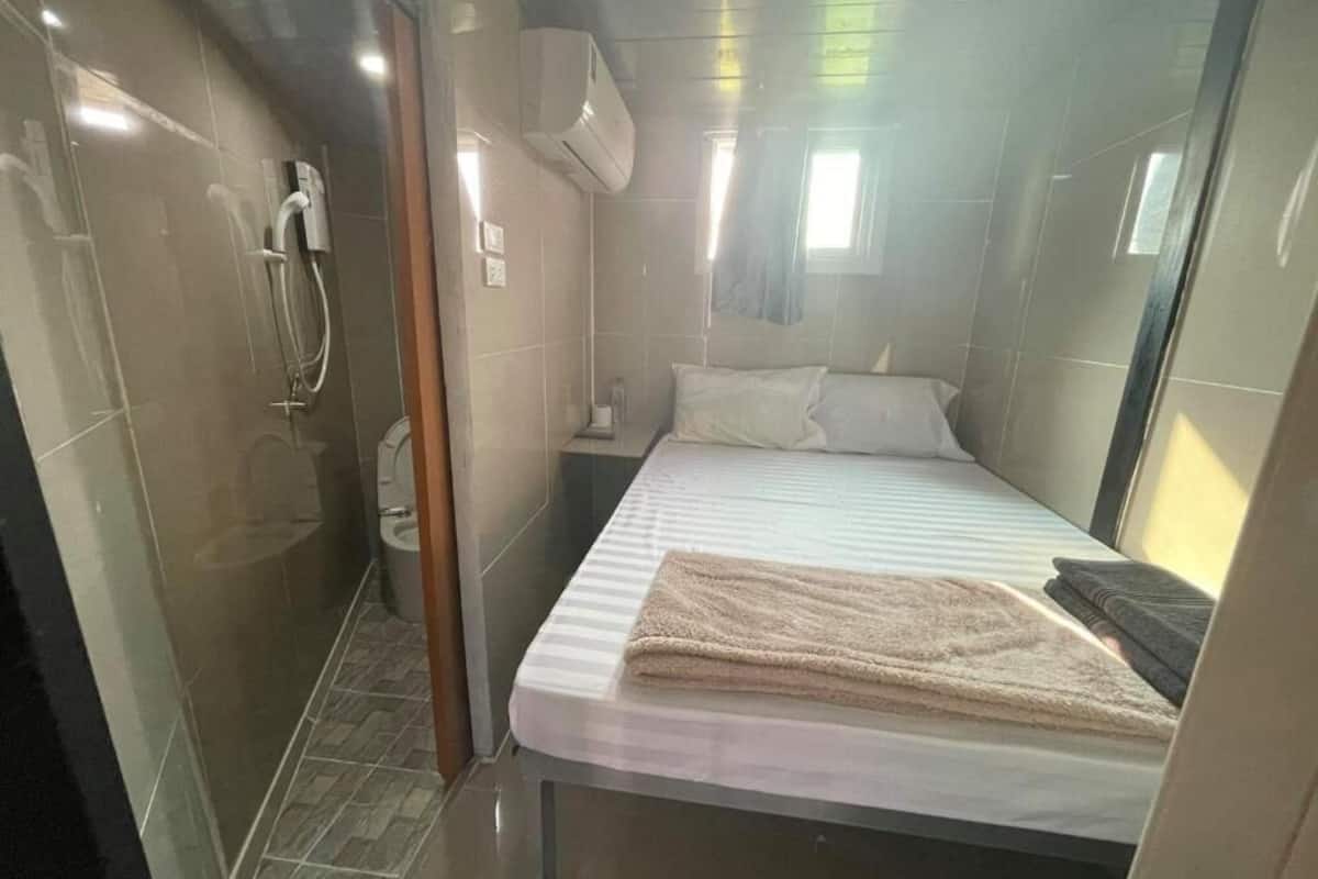 Habitación de hotel compacta con ducha, cama con colchón blanco y manta color beige, y una ventana pequeña que ofrece luz natural.