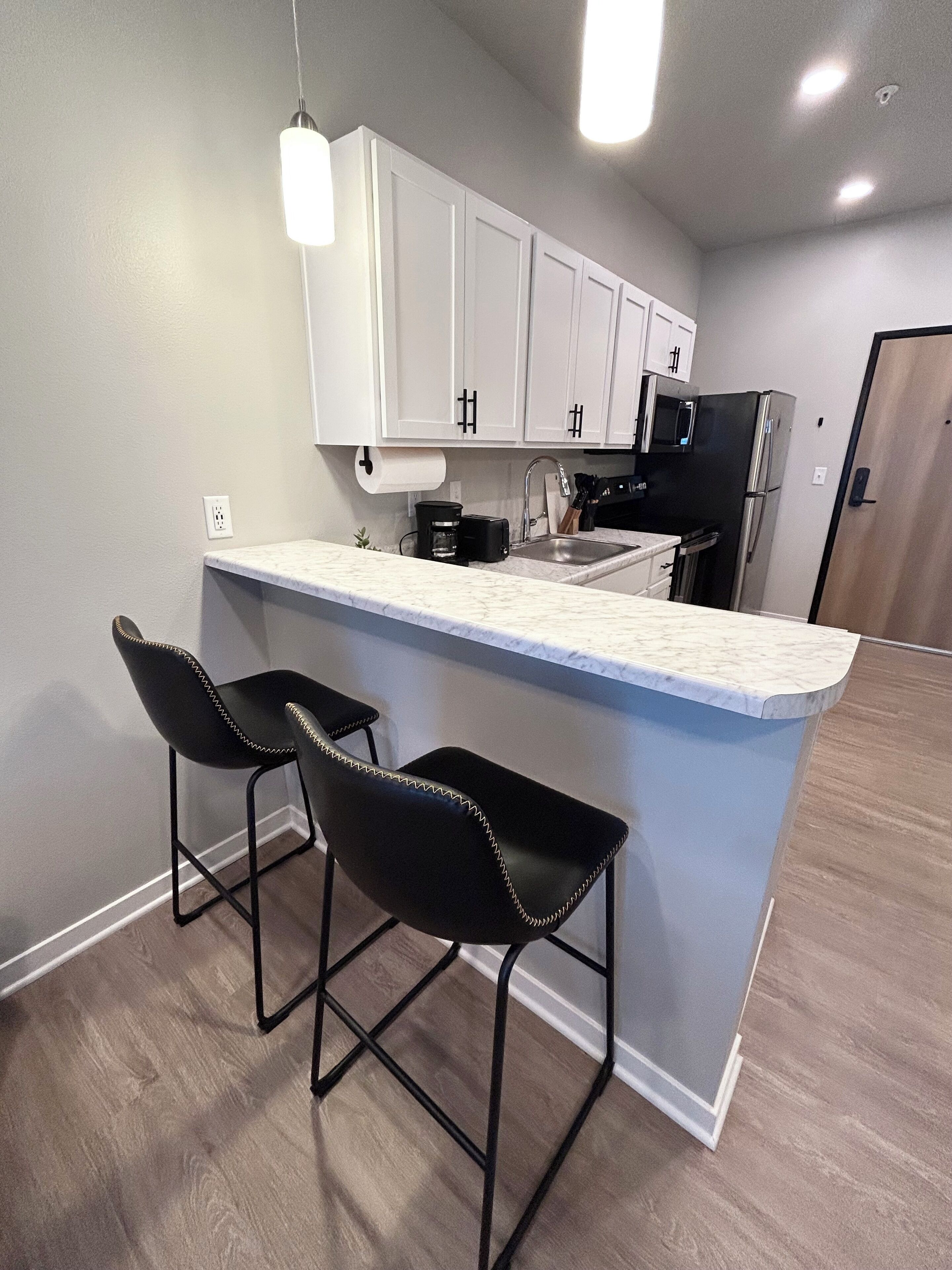 A modern kitchen with white cabinets, a marble countertop, two black bar stools, and a black refrigerator.