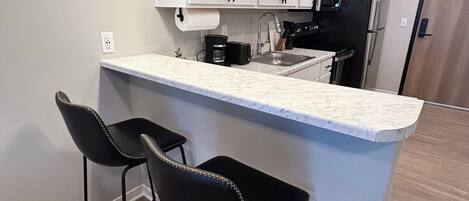 A modern kitchen with white cabinets, a marble countertop, two black bar stools, and a black refrigerator.