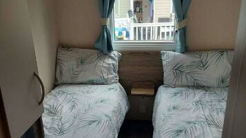 A small, enclosed room with two single beds, a window with curtains, and a view of a house outside.