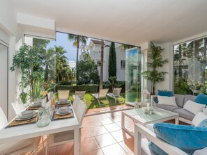 A modern outdoor dining area with a glass table and chairs, surrounded by lush greenery and a view of a residential area. - Charming 2-bedroom Lagoon Apartment (Casares)