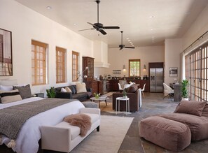 Uma sala ampla com uma cama grande, um sofá, uma mesa de centro e uma mesa de jantar. Dá para ver uma cozinha ao fundo, com uma geladeira e um micro-ondas.