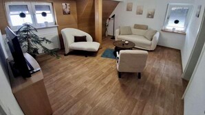 Una sala de estar con un sofá, dos sillones, una mesita de centro y un televisor. Hay dos ventanas con persianas, una planta y un radiador.